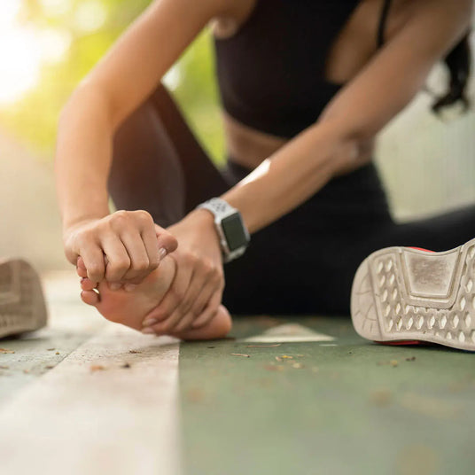 A close-up lifestyle shot of a woman in a black sports bra and leggings stretching her foot outdoors, wearing a silver smartwatch. Her white athletic shoe sole is visible in the foreground on a green surface.