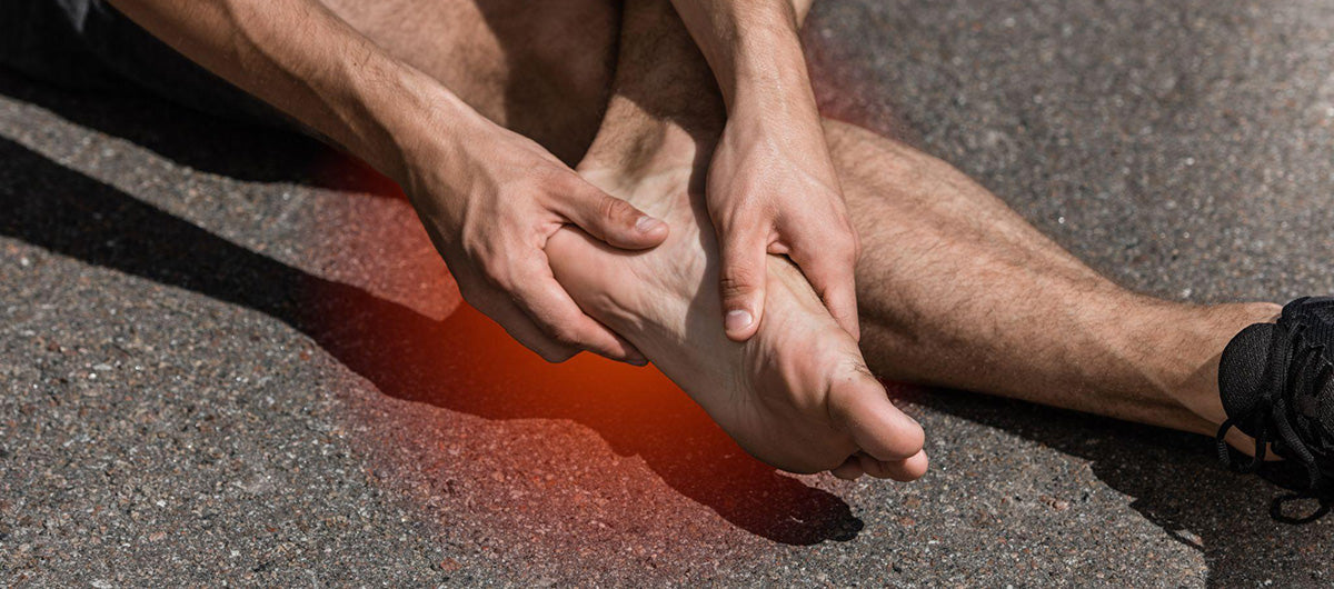 Man sitting down in shorts on asphalt rubbing heel and bottom of foot.