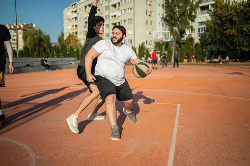 Two men playing basketball on an outdoor court, with one in a white t-shirt and black shorts dribbling a ball while being defended by another player in a dark shirt and shorts. Apartment buildings and trees are visible in the background.